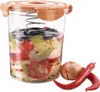 A clear glass container filled with layered vegetables and spices, with a spring mechanism inside, sits next to an onion and two red chili peppers. Anteprima dell’immagine prodotto