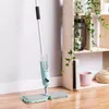 A spray mop is shown standing on a wooden floor next to a white wall and a wooden shelf unit with plants and decorative items. Náhled obrázku produktu