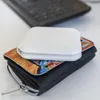 A white wireless charger is placed on top of a wallet, which is on a white surface. A laptop keyboard is visible in the background. Produktbild-Vorschau