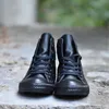 A pair of black high-top Converse sneakers are shown from a front-facing perspective, resting on a textured concrete surface with a blurred green background. Produktbild-Vorschau