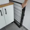 A hand is shown folding a black metal drying rack next to a white cabinet. Náhľad obrázku produktu