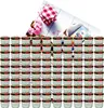 A large collection of small glass jars with red and green lids, arranged in rows, with a decorative checkered lid and pink flowers in the background. Voir les images du produit