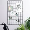 A black metal grid photo frame mounted on a wall, decorated with several small photos and a motivational quote card. A black desk lamp and some green plants are visible in the foreground. Anteprima dell’immagine prodotto