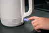 A close-up shot of a person's finger pressing the power button on a white electric kettle. Voir les images du produit