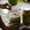 A person is placing a red rubber seal onto a glass jar filled with a light-colored liquid. Another glass jar filled with pickled items is visible in the background. Anteprima dell’immagine prodotto