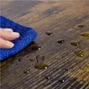 A hand holding a blue microfiber cloth wiping water droplets off a dark wood-grain surface. Produktbild-Vorschau