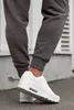 A close-up shot of a person's legs wearing white sneakers and dark gray sweatpants, standing on a paved surface next to a textured wall. Produktbild-Vorschau