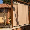 An outdoor scene with a beige curtain hanging from a pergola, with a building and plants in the background. Produktbild-Vorschau