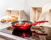 A red frying pan with tomatoes sits on a black induction stovetop, with bread and wooden crates in the background. Produktbild-Vorschau