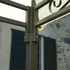 A close-up view of the metal frame of a garden pavilion, with a stone building and shutters in the background. Produktbild-Vorschau