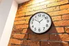 A round clock with a black frame is mounted on a brick wall. The clock has white hands and black numbers indicating the time. Anteprima dell’immagine prodotto