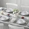 A dining table set for four people with white plates, bowls, cutlery, candles, and a floral centerpiece. Produktbild-Vorschau