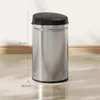 A stainless steel sensor trash can with a black lid is shown against a light-colored wall and wooden floor. Produktbild-Vorschau