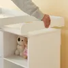 A person's hand is shown adjusting a removable changing pad on a white changing table. Two stuffed animals are visible in a shelf below. Podgląd zdjęcia produktu