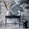 A black console table with a ribbed wood-look finish stands against a black and white forest-themed wallpaper. Dried pampas grass in a white vase sits on the table, along with decorative items. A black stool with books is placed next to the table. Produktbild-Vorschau