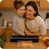 A mother and daughter are in a kitchen, smiling and looking at a vacuum sealer with a piece of salmon inside. Podgląd zdjęcia produktu