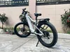 A white electric mountain bike is parked on a paved surface next to a building with some plants. Produktbild-Vorschau