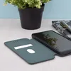 A dark green flip-style phone case is open on a light-colored wooden surface, next to a smartphone. A potted plant and a notebook with a pen are in the background. Produktbild-Vorschau