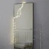 A tall mirror with fairy lights draped over it, next to a bare white tree and some wrapped gifts. Produktbild-Vorschau