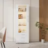 A white high-gloss display cabinet with glass doors and LED lighting, showcasing decorative items on its shelves. The cabinet has three drawers at the bottom. It is placed in a room with a chair and a potted plant nearby. Produktbild-Vorschau