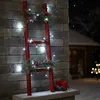 A red ladder decorated with pine branches, red berries, and white LED lights, standing against a stone wall in a snowy outdoor setting at night. Náhled obrázku produktu