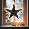 A black star-shaped lamp with small cutouts is placed on a windowsill during winter. The background shows a snowy scene with blurred lights. Produktbild-Vorschau