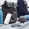 A white Bluetooth speaker is attached to a black backpack, with a blue water bottle and sunglasses nearby. A person is sitting in the background. Anteprima dell’immagine prodotto