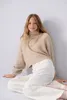 A young girl wearing a beige turtleneck sweater and white pants sits on the floor against a light gray wall. Produktbild-Vorschau