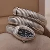 A close-up shot of a silver snake-shaped watch with a black face, resting on a beige surface. Náhled obrázku produktu