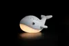 A white whale-shaped night light is illuminated from within, casting a warm yellow glow. Produktbild-Vorschau