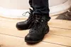 A person wearing black work boots and blue jeans standing on a wooden floor. Náhled obrázku produktu