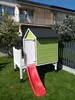 A small wooden playhouse with a red slide and a white fence, set in a grassy backyard with a house in the background. Produktbild-Vorschau