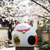 A white Maneki-neko cat figurine with a gold coin and red collar sits in front of a blurred background of cherry blossoms and traditional buildings. Produktbild-Vorschau