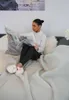 A woman is sitting on a couch reading a newspaper while wearing slippers. Produktbild-Vorschau