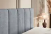 A close-up view of a gray upholstered headboard of a bed, with a bedside lamp and dried reeds in the background. Produktbild-Vorschau