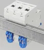 A white electrical circuit breaker is mounted on a metal DIN rail, with blue clips securing it. Produktbild-Vorschau