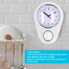 A white kitchen clock with a timer is mounted on a tiled wall, with kitchen utensils hanging below it. Náhled obrázku produktu