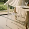 A transparent chair is placed next to a table set for a meal, with a curtain blowing in the background. Produktbild-Vorschau