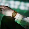A person's arm wearing a gold watch, with a blurred green background. Voir les images du produit
