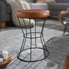 A brown leather stool with a black wire base sits on a gray rug in a living room setting. Produktbild-Vorschau