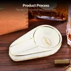 A ceramic ashtray with gold trim is placed on a wooden surface, with a cigar box and a glass of whiskey in the background. Produktbild-Vorschau