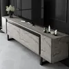 A modern TV stand with a travertine-like finish and black legs, placed in a room with a dark wall and light-colored floor. Produktbild-Vorschau