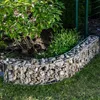 A curved gabion garden border filled with stones, separating a garden bed with plants from a lawn. Produktbild-Vorschau