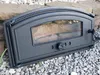 A black cast iron oven door with a glass window rests on a bed of gray gravel and large stones. Produktbild-Vorschau