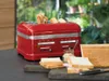 A red KitchenAid toaster with bread in it, along with slices of bread and a small jar of spread on a dark countertop. Produktbild-Vorschau
