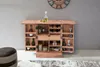 A wooden bar cabinet is open, displaying bottles, glasses, and storage compartments. It is set against a gray wall with a rug and ottoman in the foreground. Produktbild-Vorschau