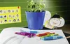 A collection of colorful pens and a pencil sharpener are laid out on an open notebook, with a potted plant and child-like drawings in the background, suggesting a classroom or study environment. Produktbild-Vorschau