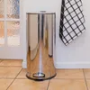 A stainless steel trash can with a foot pedal is shown in a kitchen setting, next to a door with frosted glass and a checkered towel hanging on the wall. Produktbild-Vorschau