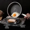A hand holding a frying pan with a fried egg in it, with a slice of toast and ham on a plate in the foreground. Náhľad obrázku produktu