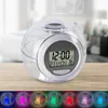 A digital alarm clock with a clear spherical casing sits on a wooden surface, with a blurred bedroom background. Below the clock, a row of smaller spherical objects are illuminated with different colors. Produktbild-Vorschau